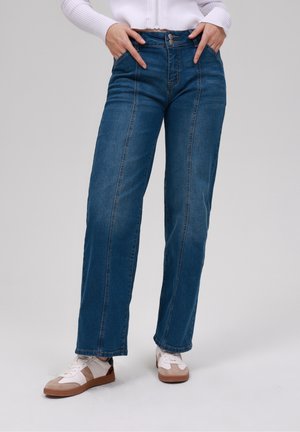 Person iført højtaljede blå jeans med forreste sømme, hænder i lommerne, hvid lynlåstrøje og hvide og beige sneakers på en ensfarvet baggrund.