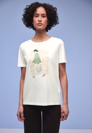 Kvinde med krøllet hår iført hvid t-shirt med illustration af en gående person i grøn top, beige bukser, sandaler og bærende en taske.