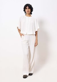 Witte, gestructureerde cropped top met een ronde halslijn en knoopsluiting, gecombineerd met losse, lichte broek en schoenen met dierenprint.