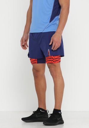 Mand iført blå atletikshorts med røde mønstrede kompressionsleggings under og sorte løbesko, stående sideværts.