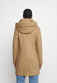 Kvinde med hår i en knold iført guldringørelser, en tan uldfrakke med bred krave, og blå jeans, vendende væk mod hvid baggrund.