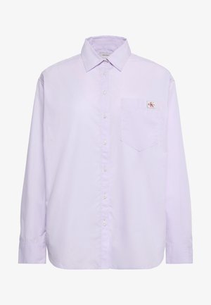 Cămașă lungă cu mâneci lungi, de culoare mov deschis, cu nasturi și buzunar pe partea stângă a pieptului, având o mică etichetă cu logo-ul Calvin Klein deasupra buzunarului.