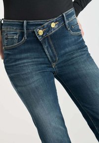 Jeans en denim bleu foncé avec une fermeture à double bouton, un design à cinq poches et des détails de couture contrastés présentant un léger effet de décoloration.