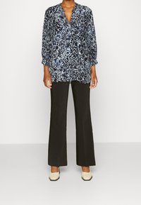 Persona vistiendo una blusa suelta de patrón blanco y negro, pantalones de pierna ancha negros y zapatos beige de punta cuadrada, de pie contra un fondo liso.