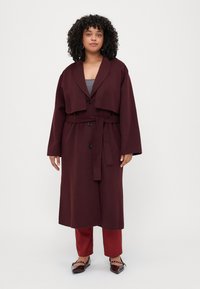 Burgundy long wool coat with a draped collar, button closure, and a matching belt, paired with red trousers and patent shoes.