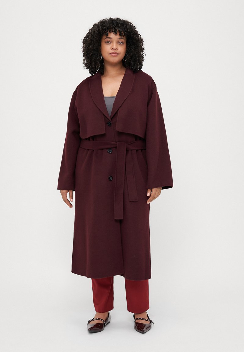 Burgundy long wool coat with a draped collar, button closure, and a matching belt, paired with red trousers and patent shoes.