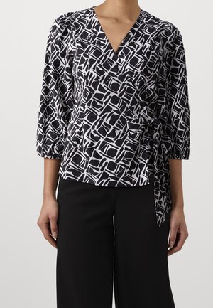 Femme portant un chemisier cache-cœur à motif géométrique noir et blanc avec manches trois-quarts et un pantalon noir à jambes larges.