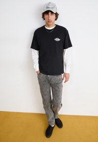 Camiseta negra de manga corta superpuesta sobre una camiseta blanca de manga larga, pantalones de mezclilla gris, zapatos negros y una gorra gris con detalles de logo.