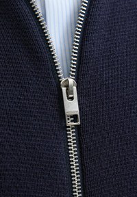 Jūrinis zip-up drabužis su tekstūruotu mezgimo paviršiumi. Yra sidabrinė metalinė užtrauktukas su firminiu rankenėle ir šviesiai mėlynos juostelės pamušalas.