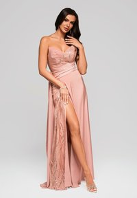 Nainen strapless, paljetein koristellussa vaaleanpunaisessa mekossa, jossa on reiden ylle ulottuva halkio, poseeraa elegantisti yksi käsi kasvojensa lähellä, neutraalin taustan edessä.