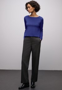 Haut violet à manches longues avec un col rond, associé à un pantalon gris taille haute. Mannequin portant des bottines noires. Fond uni.