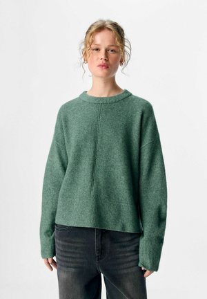 Jonge vrouw met krullend haar, gekleed in een losse groene trui en versleten zwarte jeans, tegen een effen achtergrond.