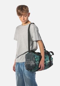 Green and black duffel bag with a rounded shape, made of a glossy material; features a logo and black straps for carrying.