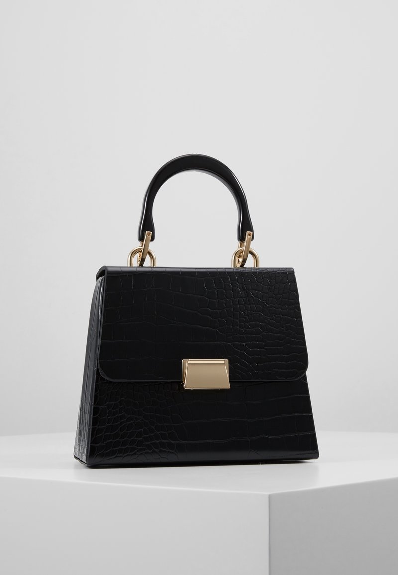 Sac à main texturé noir avec une forme structurée, un fermoir rectangulaire doré et une poignée noire solide avec des ferrures dorées sur une surface blanche.