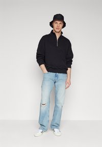 Joven con un sombrero tipo cubo negro, sudadera negra con media cremallera, jeans claros desgastados y zapatillas blancas con rayas negras, de pie contra una pared lisa.