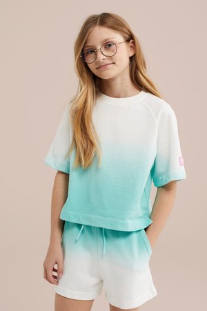Jong meisje met lang blond haar, ronde bril en een wit-turquoise ombre kortemouwen top en bijpassende korte broek, staand tegen een beige achtergrond.