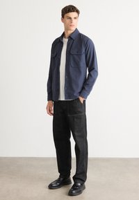 Mørkeblå skjorte med knapper og to brystlommer, brukt over en hvit T-skjorte, kombinert med mørke jeans og svarte slip-on sko. Jevn stofftekstur.