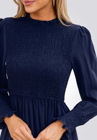 Vestido de marina con cuerpo fruncido, mangas largas abullonadas y puños con volantes. Material suave con sección superior texturizada. Ajustado en la cintura.