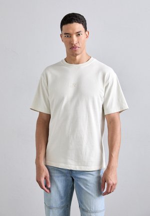 Short-sleeved cream T-shirt in soft cotton with a round neck and subtle embroidered logo on the chest, paired with light blue denim shorts.