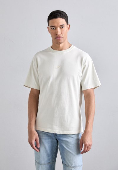 Tricou cu mânecă scurtă, de culoare crem, fabricat din bumbac moale, cu guler rotund și un logo brodat subtil pe piept, asortat cu pantaloni scurți din denim de culoare albastru deschis.