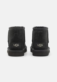 Bottines en suédine noire avec une doublure douce, présentant un petit patch logo UGG, des coutures texturées et une semelle en caoutchouc plate.