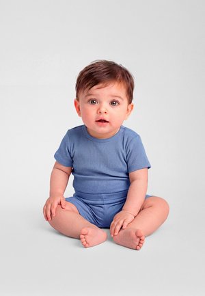 Beebi pruunide juuste ja suurte silmadega, istub jalad ristis hallil põrandal, seljas sinine lühikese varrukaga soonikust kombinesoon, vaatab otse ette, suu kergelt avatud.