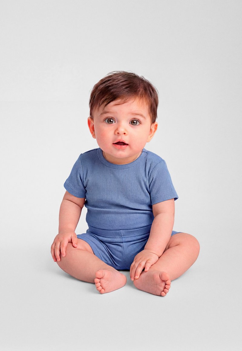 Bébé aux cheveux bruns et aux grands yeux assis en tailleur sur un sol gris, portant une tenue côtelée à manches courtes bleue, regardant droit devant avec la bouche légèrement ouverte.