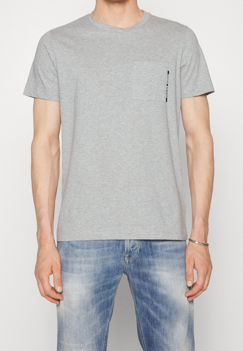 T-shirt en coton gris à manches courtes, col rond et poche avant. Présente un petit logo imprimé près de la poche.