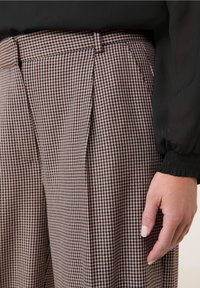 Rutiga byxor i beige och bruna nyanser, med hög midja, veck och en mjuk tygstruktur. En hand syns vila på byxorna.