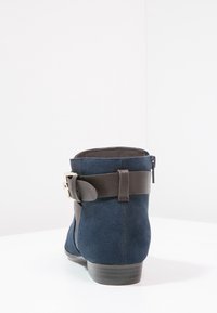 Mockasins med ankeldesign i marinblått med en rem i brunt läder och spänne. Har en låg klack och dragkedja på sidan.