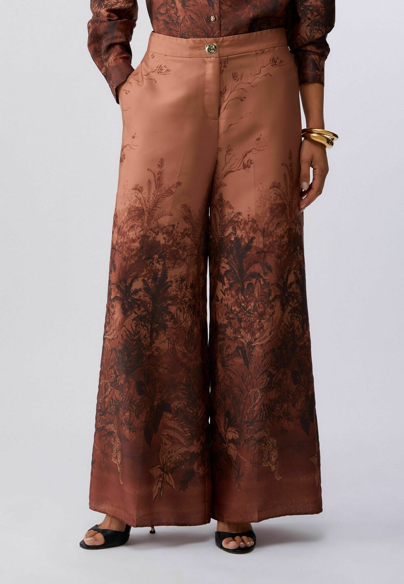 Pantalon large marron à imprimé botanique foncé, avec une légère brillance, taille haute, associé à des talons ouverts noirs et des bracelets dorés.