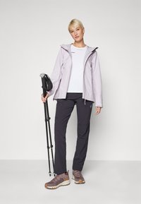 Light purple waterproof jacket with a hood, white shirt underneath, dark pants, and brown trail shoes. Holding a black trekking pole.