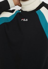 Zwarte trui met turkooizen en crèmekleurige schouderdetails, geribde hals en geborduurd "FILA"-logo in wit en rood op de borst.