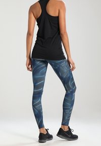 Donna con un top nero a spalla larga e leggings blu a fantasia, in piedi con le mani rilassate ai lati su uno sfondo semplice.