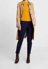 Beige trench coat med krage och stora knappar, parat med en texturerad senapsgul tröja, marinblå byxor och bruna klackskor med glänsande yta.