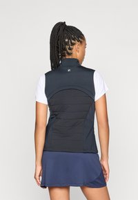 Svart ermeløs vest med høy krage, med polstret tekstur på baksiden og en logo. Kombinert med en hvit kortermet topp og et blått skjørt.