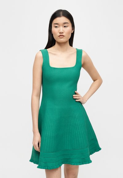 Femme aux longs cheveux noirs portant une robe en maille côtelée verte sans manches avec un décolleté carré et un ourlet à volants, debout devant un fond uni.