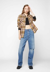 Camisa de flanela xadrez em tons de amarelo, rosa e castanho, usada sobre um top cropped preto, combinada com jeans largos de cintura alta em azul claro e botas pretas.