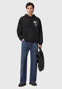 Haut à capuche noir avec un graphique de crâne, jean bleu et chaussures sombres. Tenant un sac noir, affichant une coupe décontractée et un style informel.