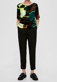 Bluzka z krótkim rękawem w czarny, zielony i brązowy abstrakcyjny wzór; zestawiona z czarnymi spodniami i masywnymi czarnymi mokasynami, tworząc spójną stylizację.