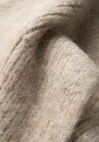 Tessuto lavorato a maglia beige con una texture morbida e pelosa. Le fibre sono intrecciate in modolasso, creando un aspetto caldo e accogliente con dettagli di cucitura visibili.