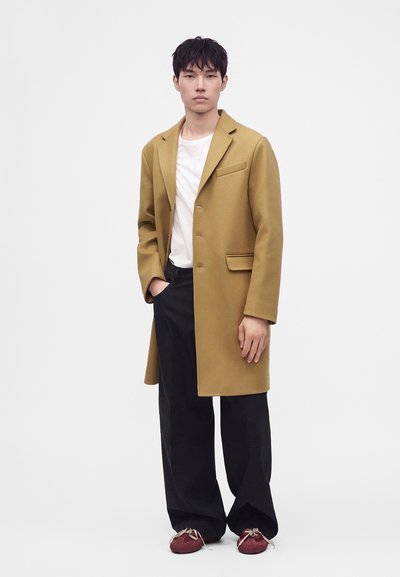 Homme portant un manteau jaune moutarde, une chemise blanche, un pantalon large noir et des chaussures rouges, debout avec une main dans la poche sur un fond uni.