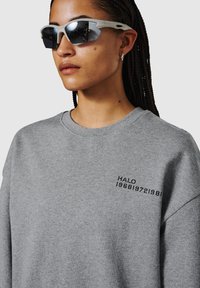 Grijze sweatshirt met een ronde halslijn, met zwarte geborduurde tekst "HALO 196819721980" op de linker borst. Model draagt reflecterende zonnebril.
