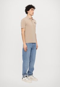 Beige kortermet polo skjorte med krage og brodert logo, parret med lyseblå jeans og hvite joggesko, stående mot en nøytral bakgrunn.