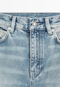 Lyseblå denimjeans i taljeområdet med forreste knap, bæltestropper, lynlås og synlige lommekanter.