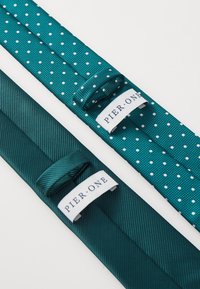 Cravatele teal au un material neted și texturat; una cu puncte albe și cealaltă într-un design solid. Ambele au etichete de marcă.
