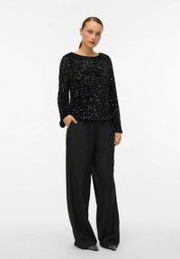 Svart paljettprydd långärmad topp med rund halsringning, ihop med högmidjade svarta vida byxor. Modellen står med en hand i fickan.