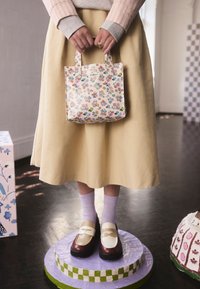 Borsa mini tote con stampa floreale, manici corti, sfondo crema e fiori colorati. Indossata con una gonna gialla e calzini viola chiaro.