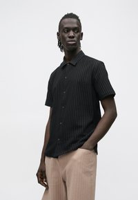 Zwart overhemd met korte mouwen en knopen, met een geribbelde textuur, voorzien van een kraag en knopen aan de voorkant, gedragen met lichtbeige textuurbroek.