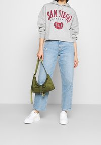 Persona che indossa un felpa grigia con la scritta "Università di San Diego", jeans azzurri, scarpe bianche, mentre tiene una borsa a spalla verde oliva su uno sfondo semplice.